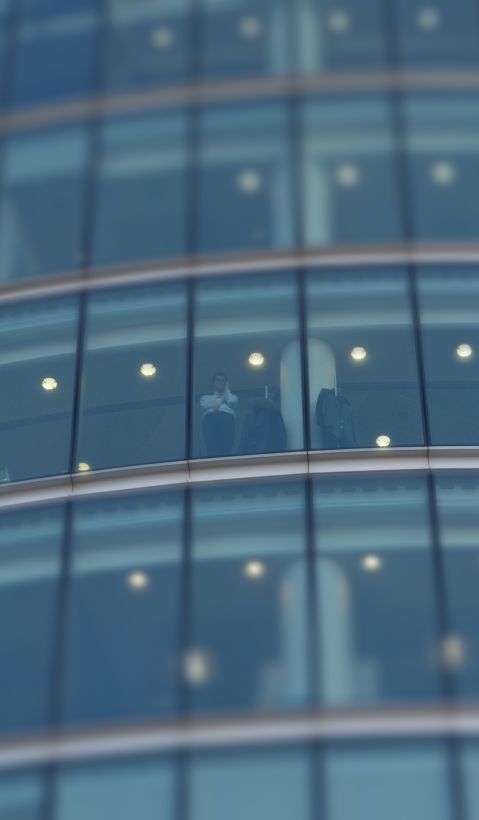 A photo of a man, standing on the window of an office building with glass facade, making a phonecall